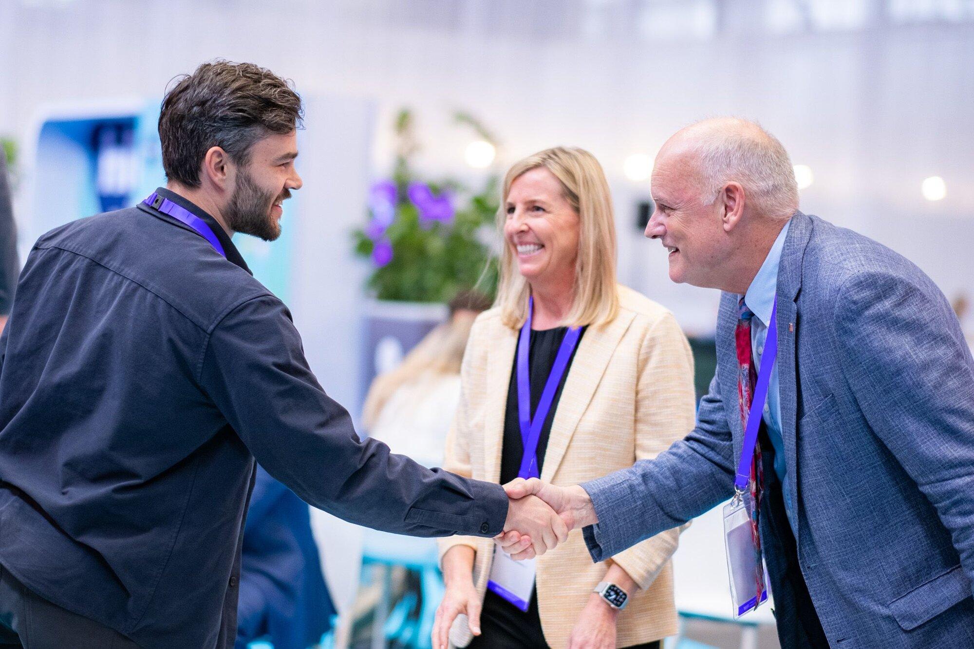 A man with a beard and dark shirt shakes hands with an older investor in a suit and tie. A woman in a beige blazer stands between them, smiling. All three wear conference lanyards, suggesting they are at a startup networking event, with blurred people and floral decor in the background.