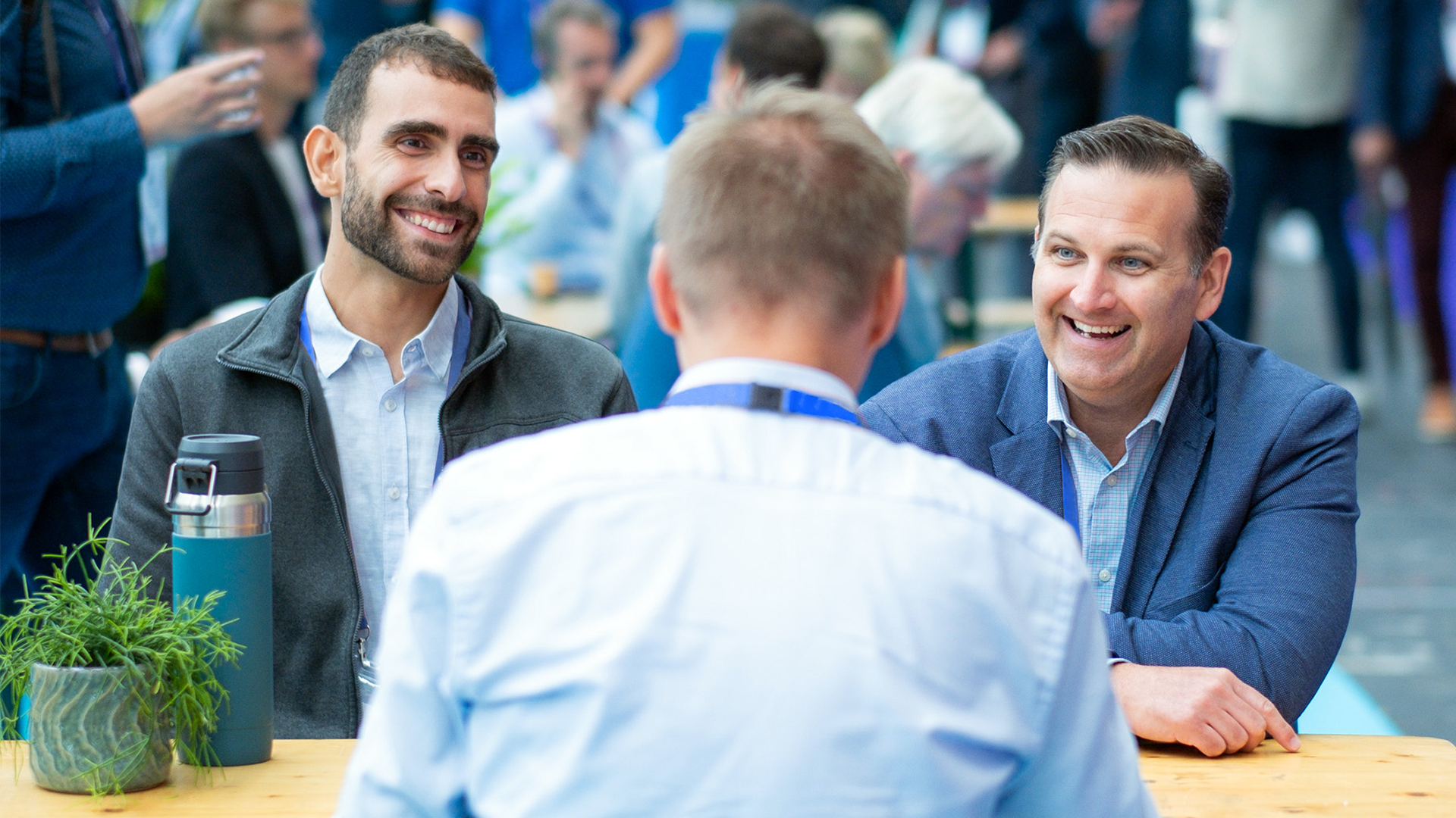 Three men are seated at a table engaged in conversation. The man on the left and the man on the right are smiling, while the back of a third man's head is visible in the foreground. A small plant and a water bottle sit on the table, creating a relaxed atmosphere perfect for an investor networking session.