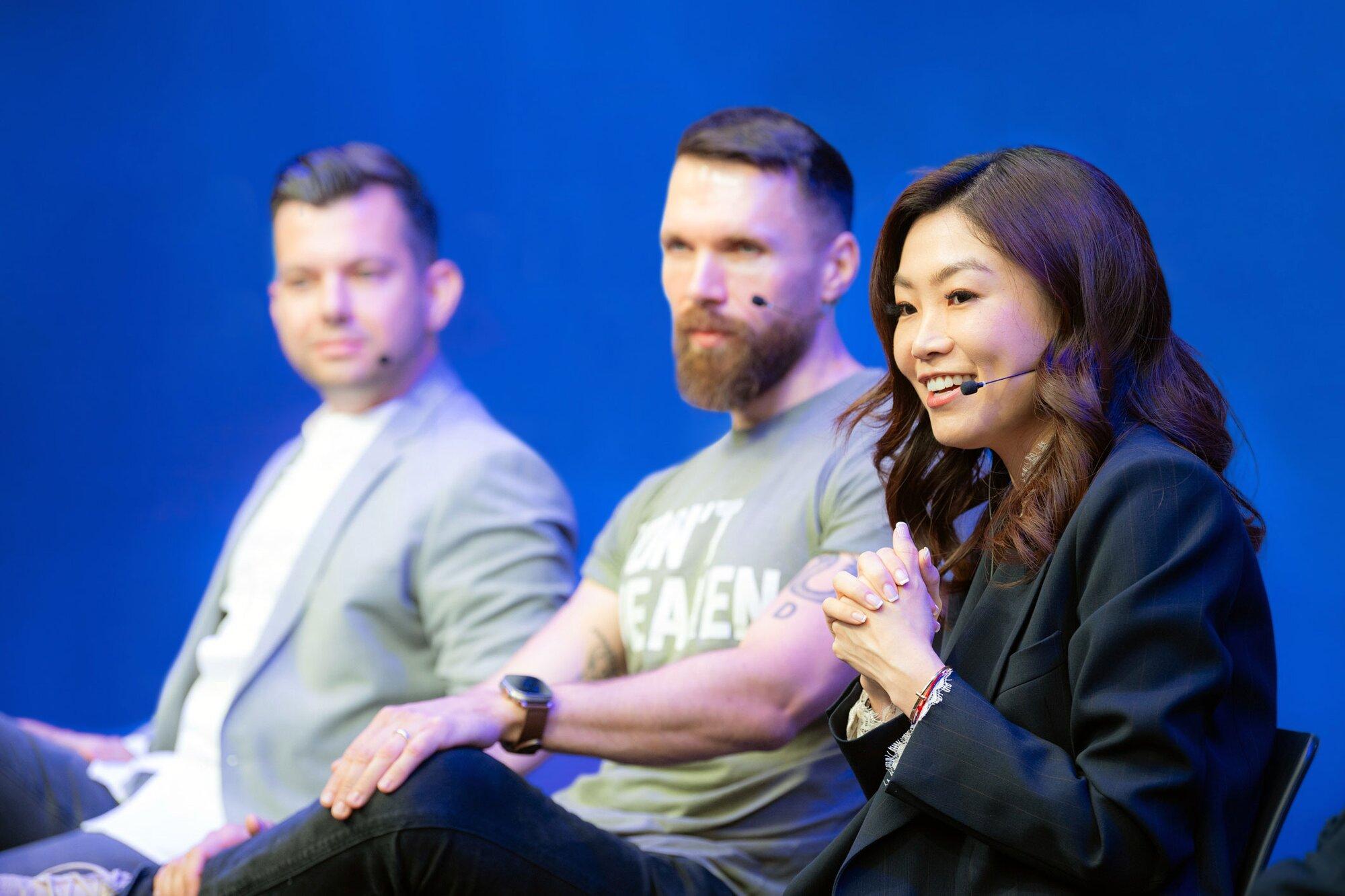 A woman with long dark hair smiles and speaks into a mic headset at the tech conference. She is wearing a dark formal jacket and has her hands clasped in front of her. A man in a t-shirt, likely representing a startup, sits beside her. The background is a solid blue color.