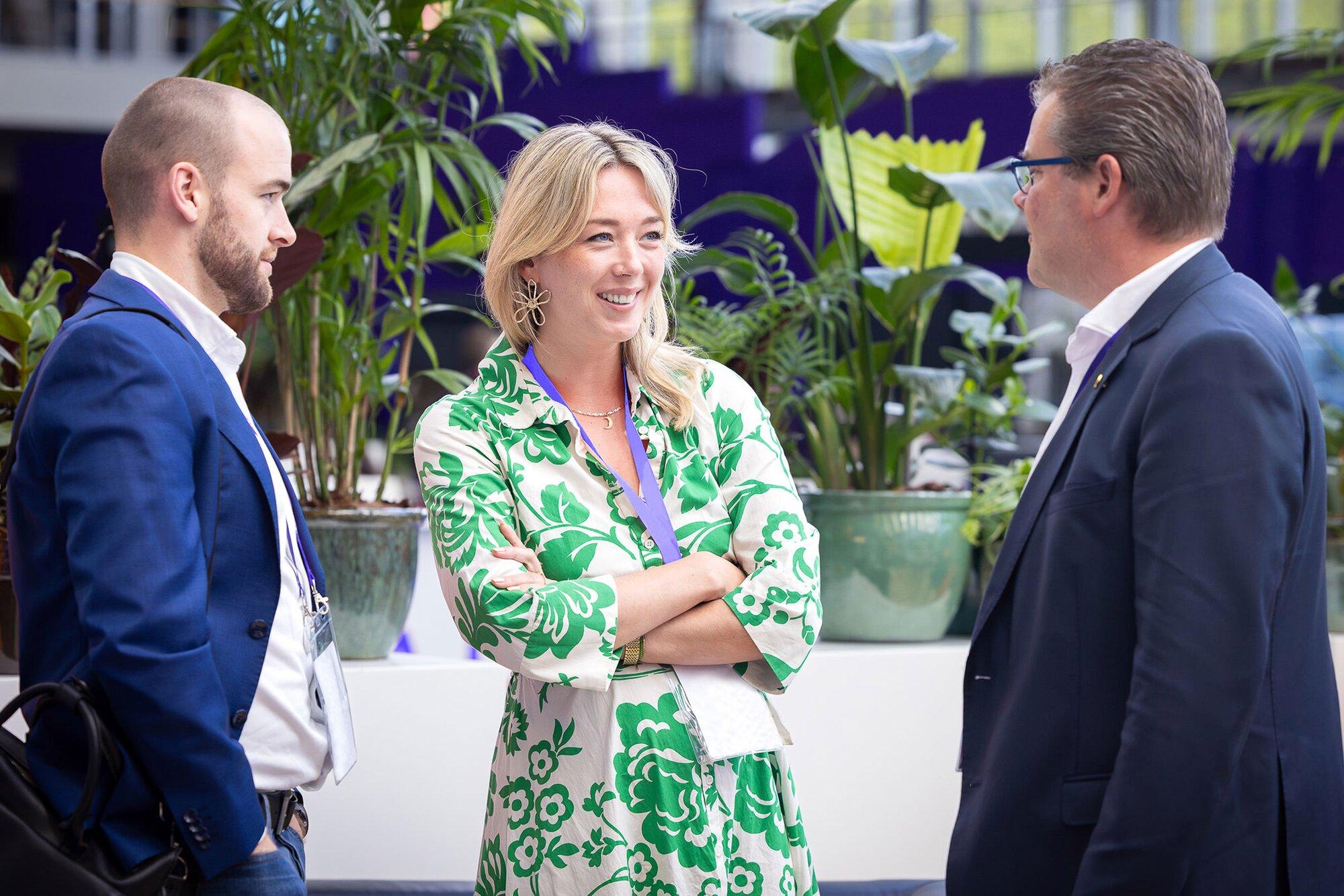 Three people are standing and engaged in conversation at a tech conference. The woman in the center is smiling, wearing a green and white floral dress. The two men, dressed in blue jackets and shirts, face her from either side. They are in a well-lit space with potted plants in the background, clearly networking.