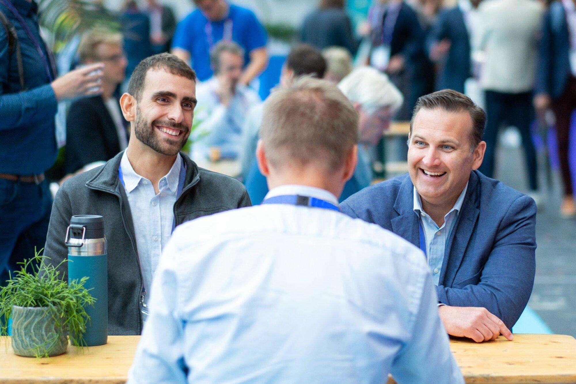 Three men are seated at a table engaged in conversation. The man on the left and the man on the right are smiling, while the back of a third man's head is visible in the foreground. A small plant and a water bottle sit on the table, creating a relaxed atmosphere perfect for an investor networking session.