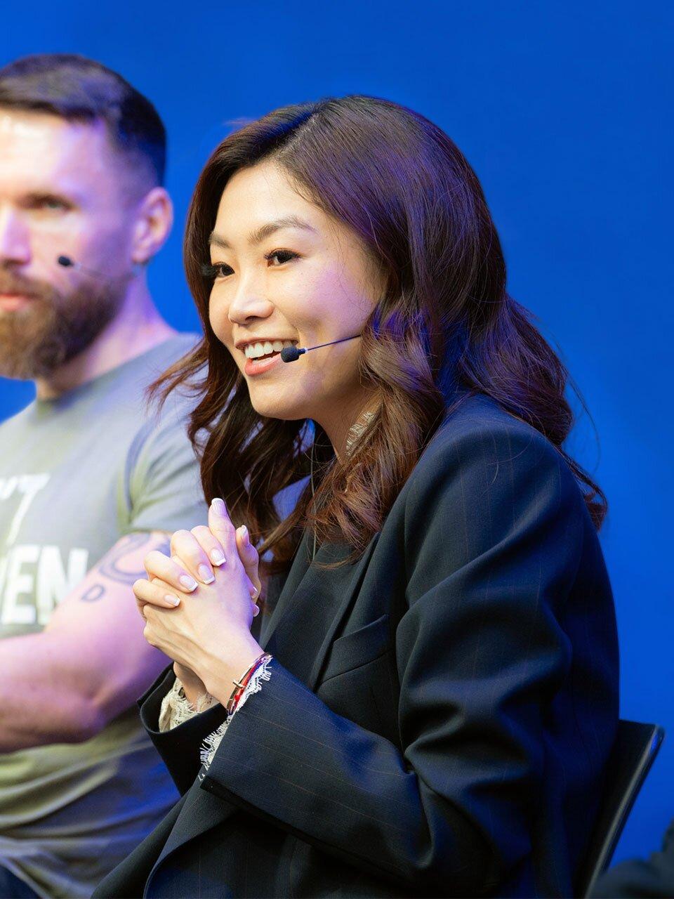 A woman with long dark hair smiles and speaks into a mic headset at the tech conference. She is wearing a dark formal jacket and has her hands clasped in front of her. A man in a t-shirt, likely representing a startup, sits beside her. The background is a solid blue color.