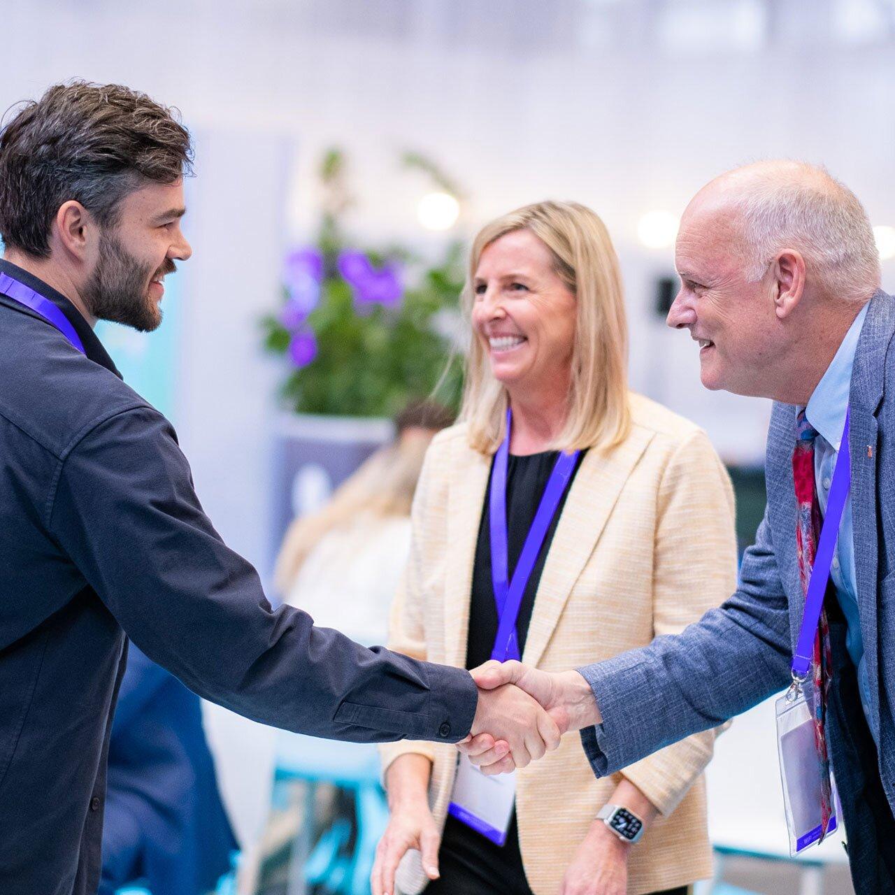 A man with a beard and dark shirt shakes hands with an older investor in a suit and tie. A woman in a beige blazer stands between them, smiling. All three wear conference lanyards, suggesting they are at a startup networking event, with blurred people and floral decor in the background.